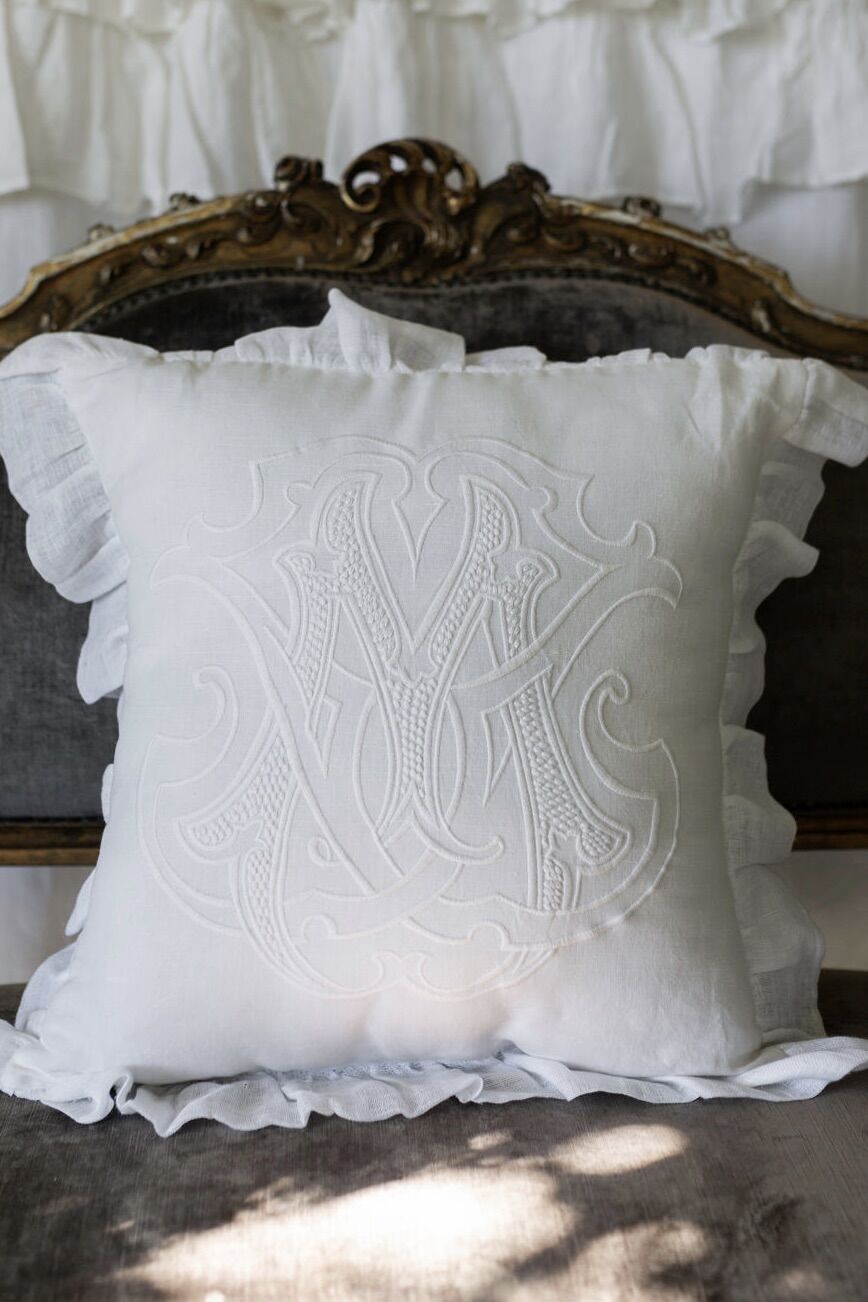 White decorative throw pillow with raised monogram embroidery on a vintage carved headboard/sofa backdrop