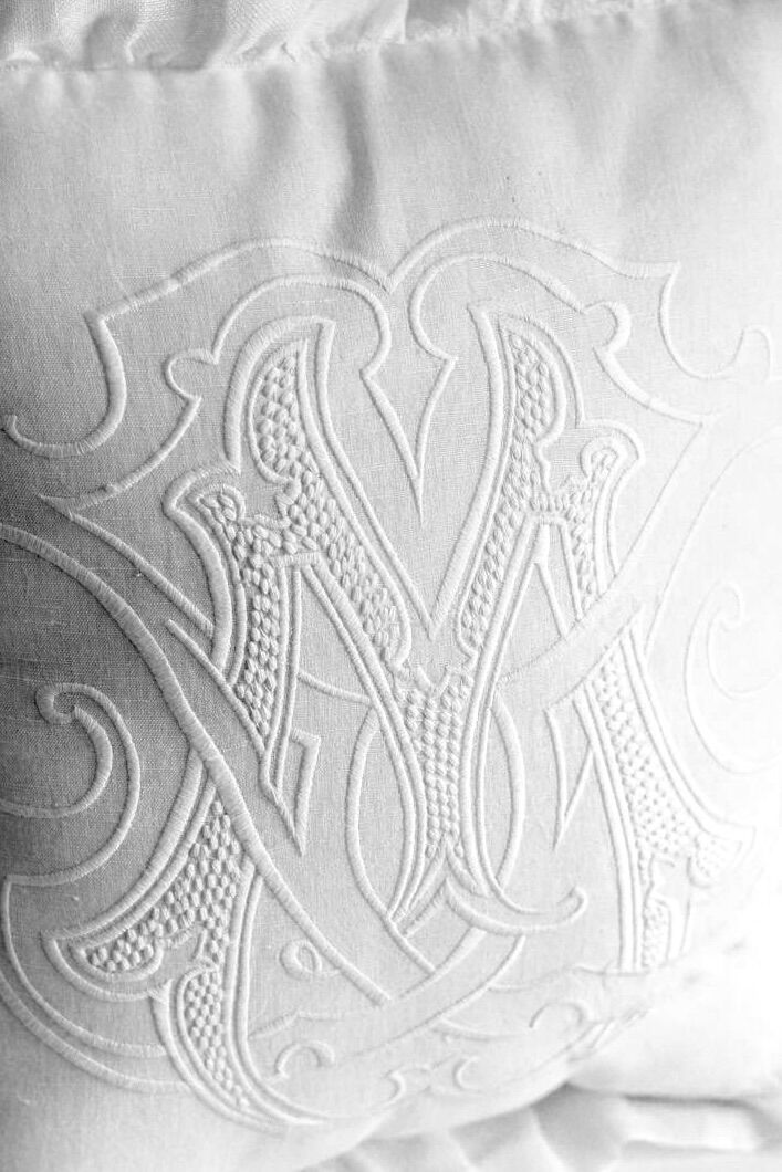 White fabric with an intricate, raised monogram of interwoven letters, decorative embroidery on a napkin or linen piece.