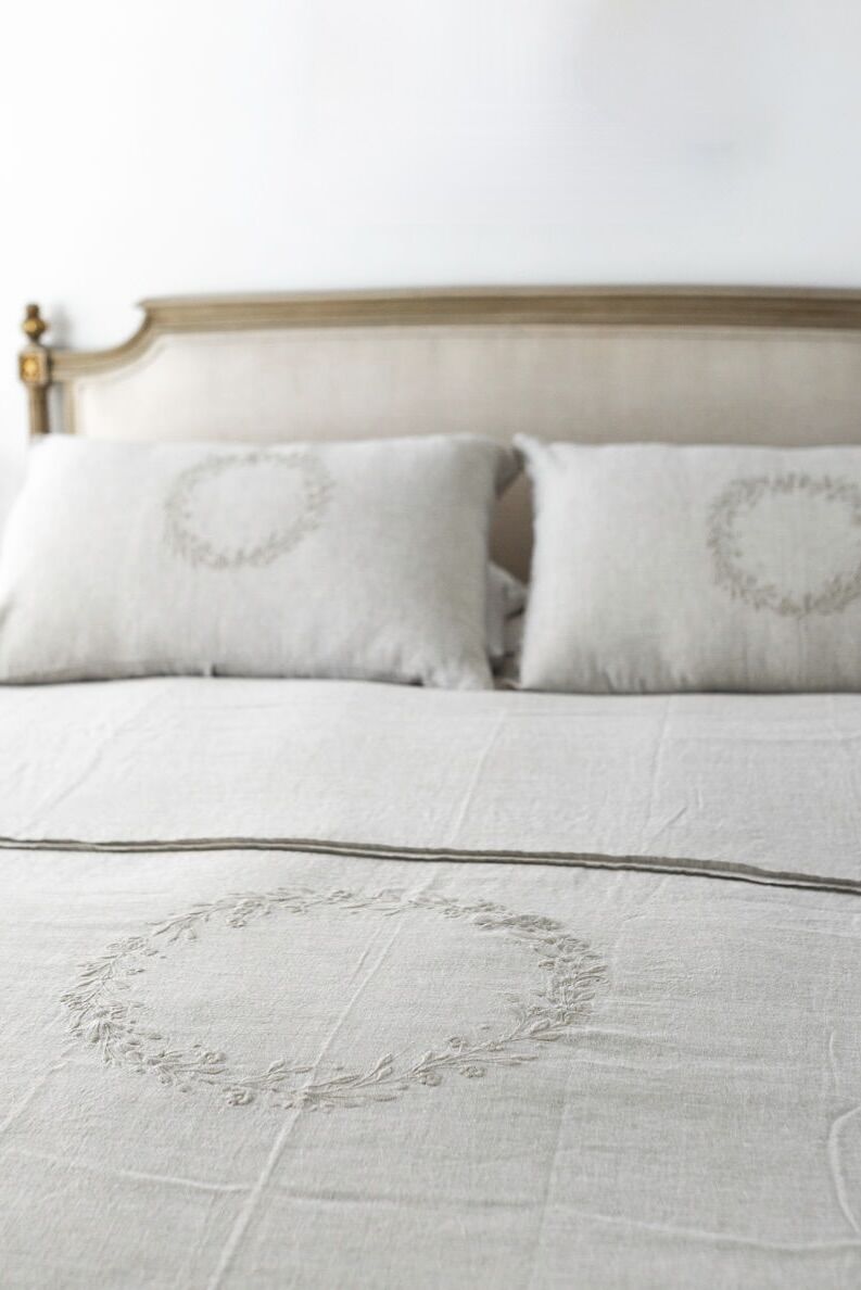Bed with cream headboard, white linens, and embroidered circular wreath designs on pillows and bedspread.