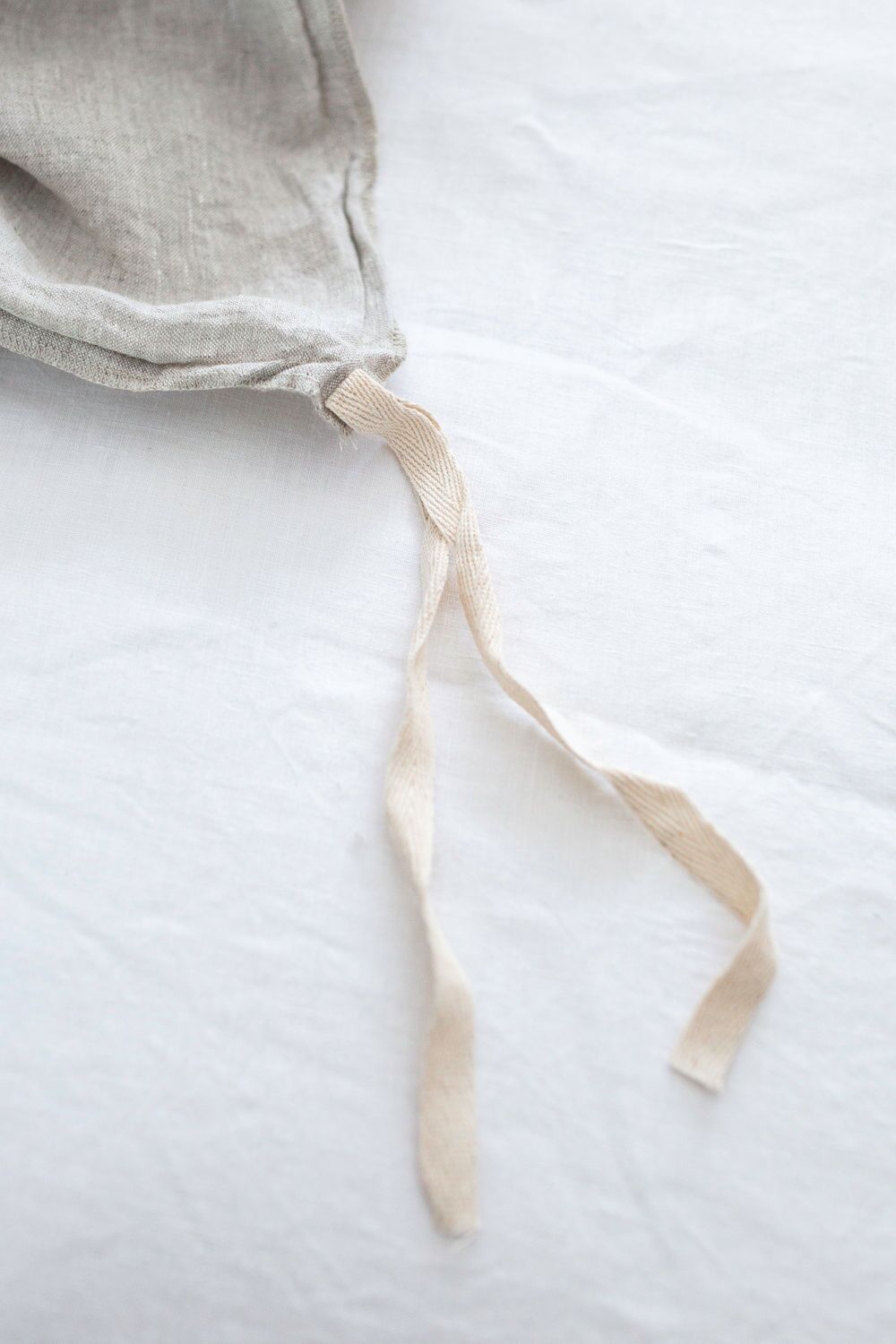 Close-up of a light linen drawstring bag resting on a white surface, with beige fabric ties hanging loose.