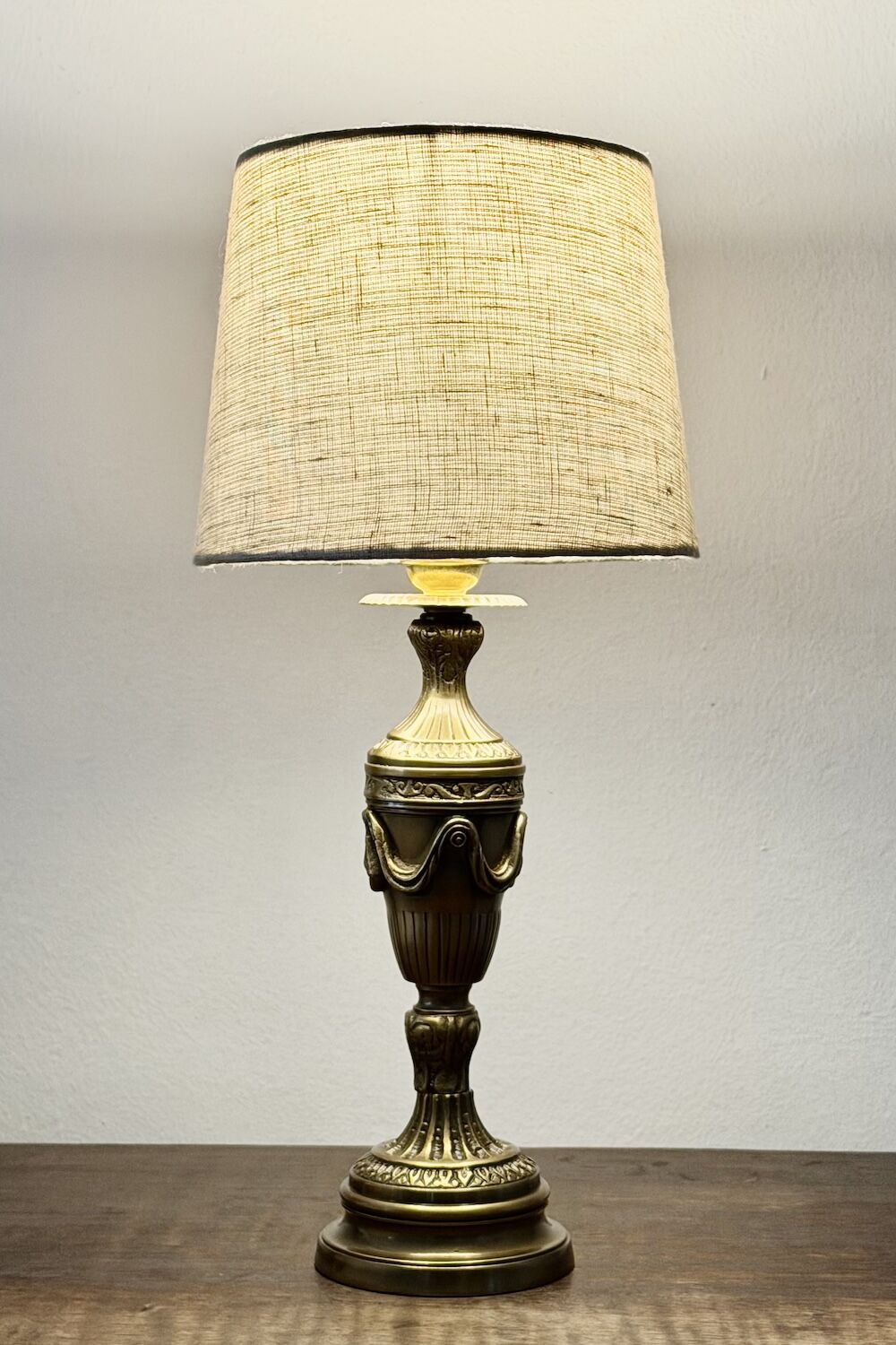 Ornate brass table lamp with a beige fabric shade on a wooden surface against a plain wall