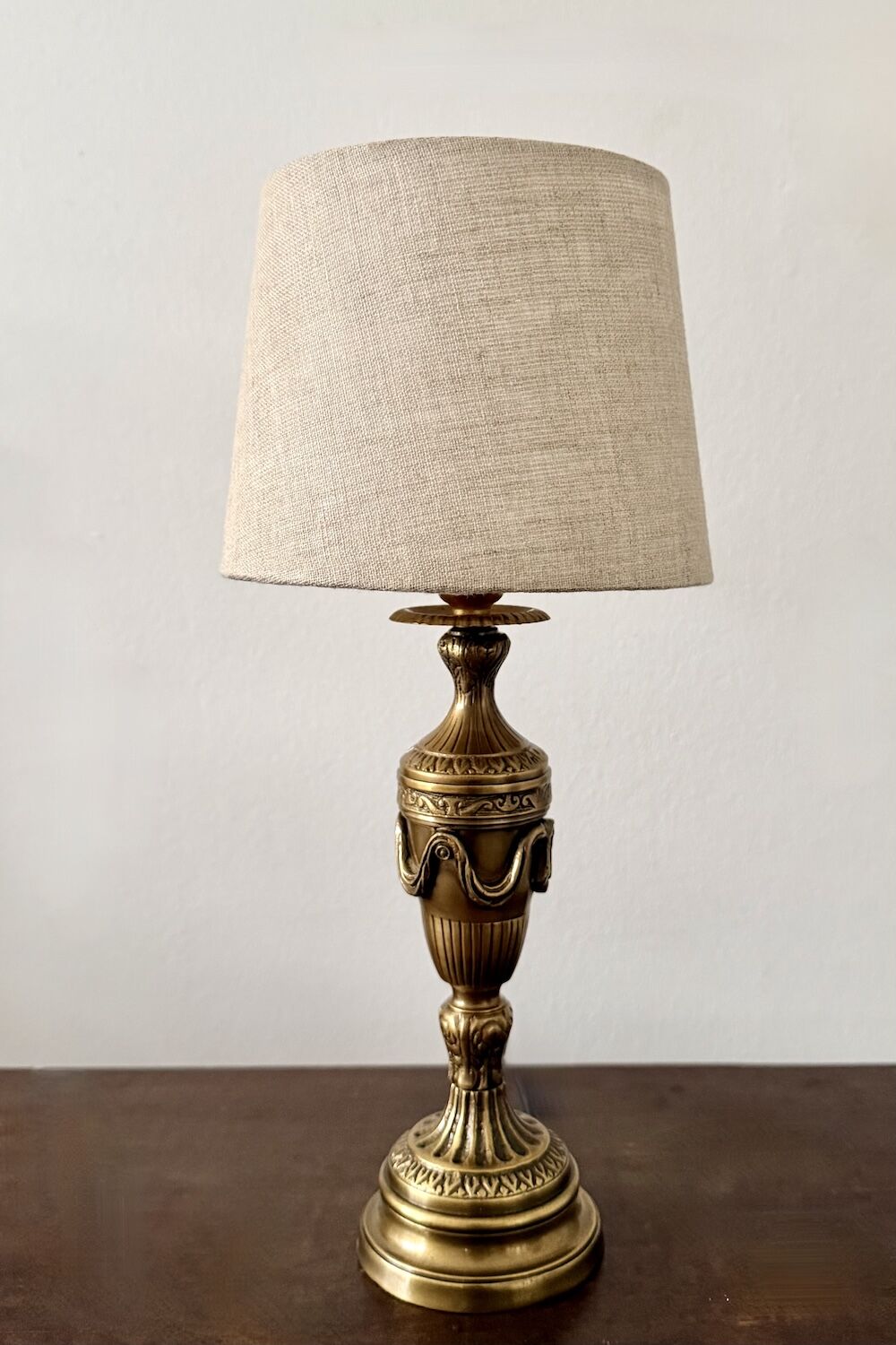 Ornate gold table lamp with a beige fabric shade on a dark wooden surface, against a light wall.