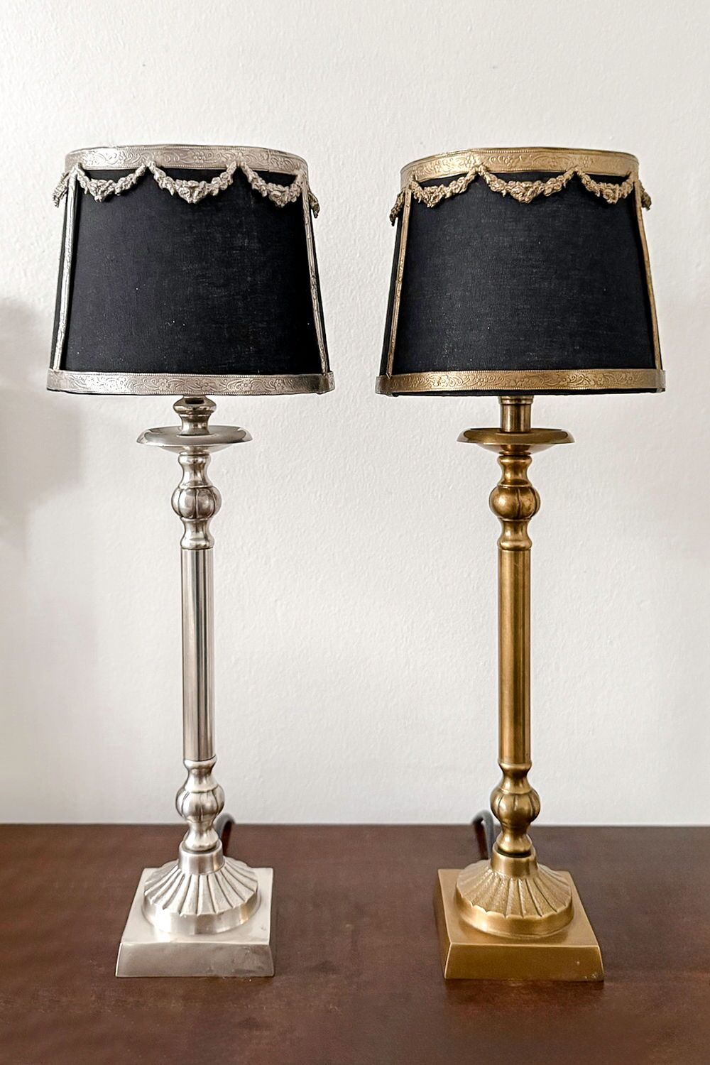 Two vintage table lamps with black fabric shades and decorative metal trim, one silver and one brass, on a wooden surface.