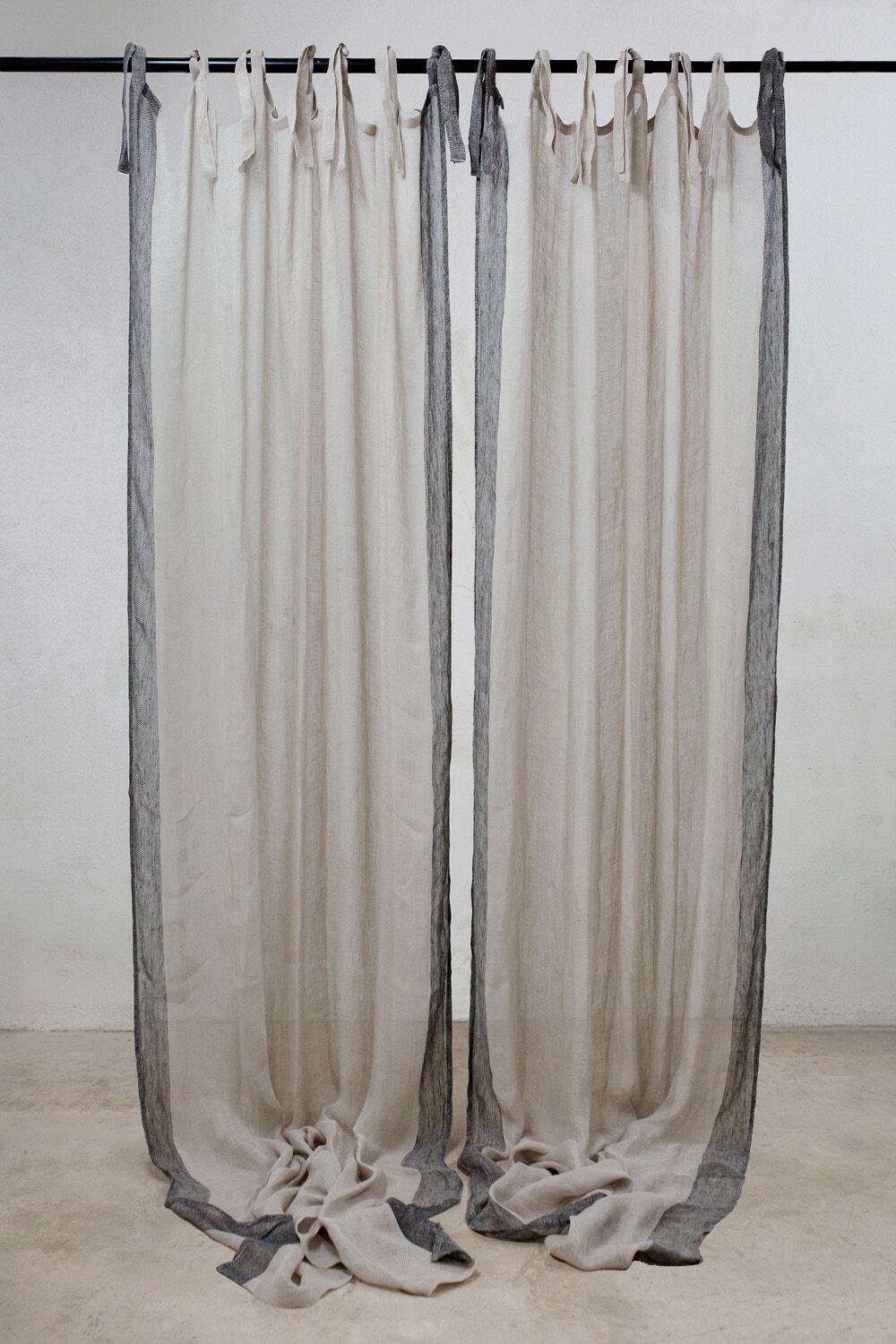 Two long beige curtains with dark gray borders hanging from a rod, pooling on the floor at the bottom.