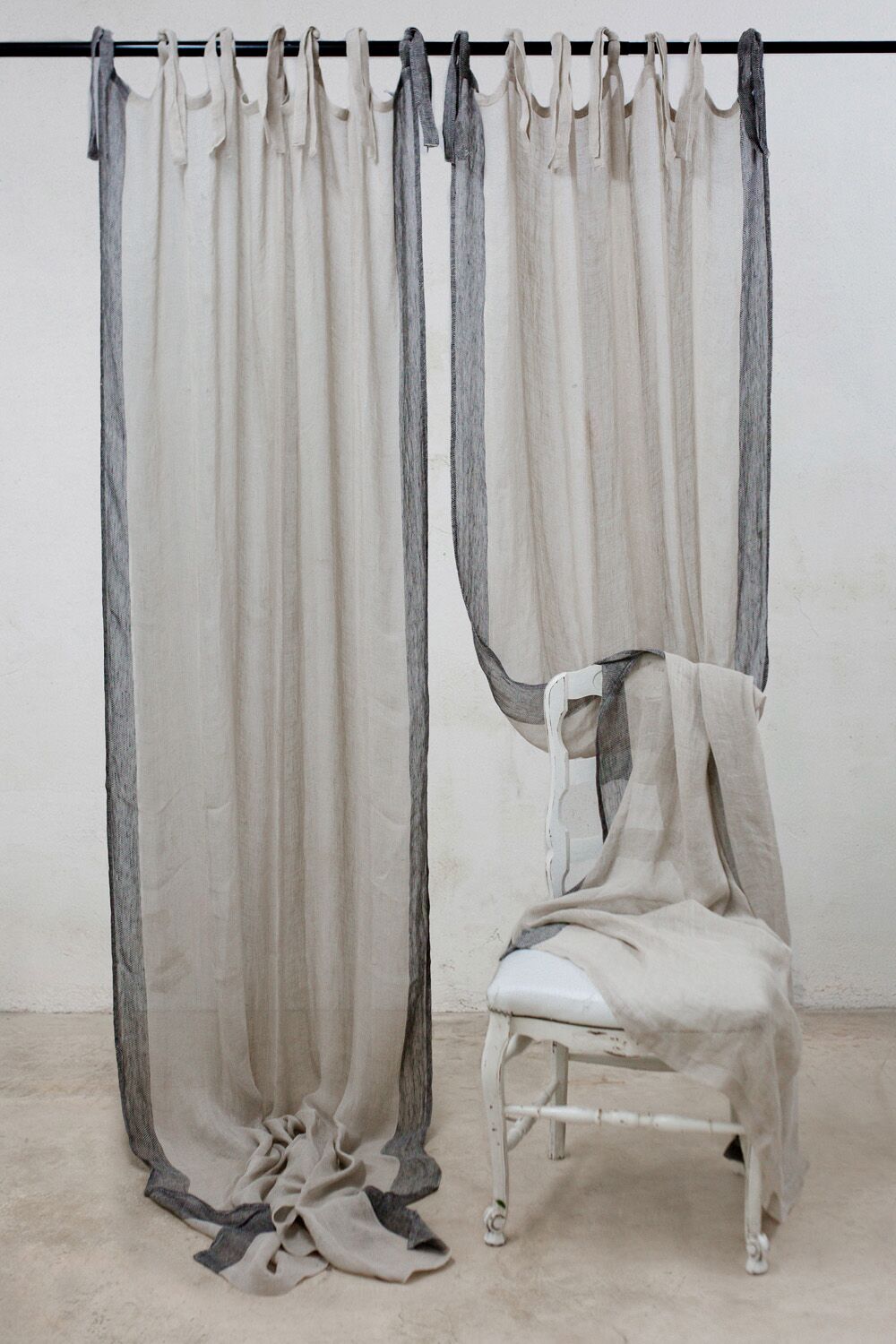 Neutral-toned fabric curtains hang from a rod, pooling on the floor, with a white chair draped in cloth beside them—an art-installation look.