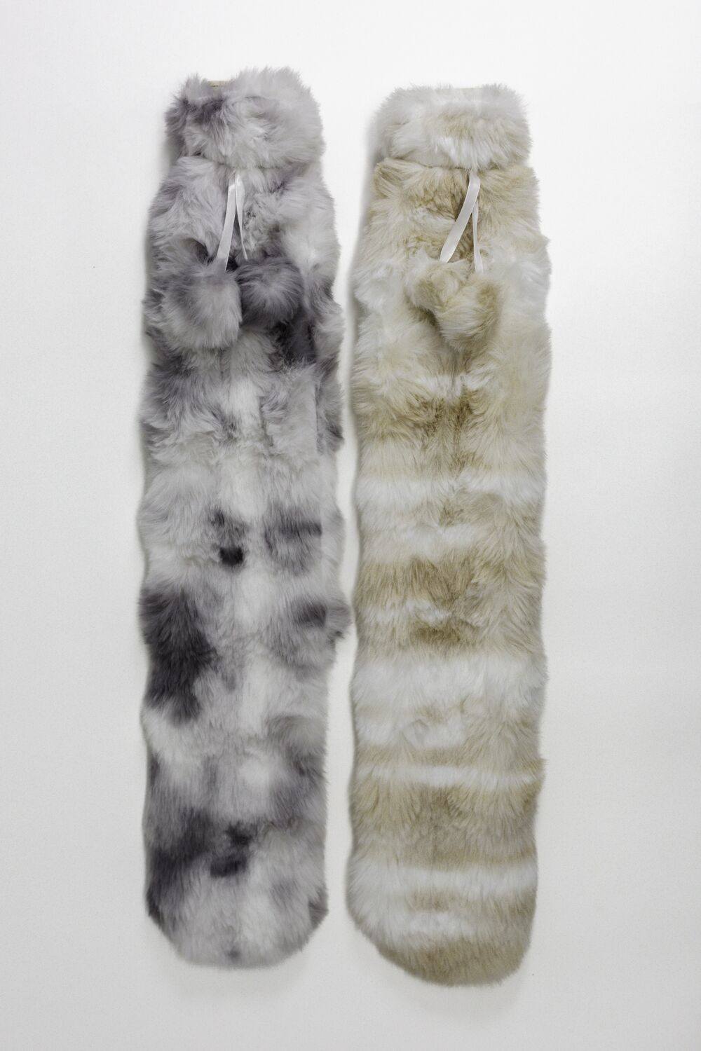 Two long faux‑fur scarves laid side by side: one gray/white, the other beige/white, each with a white ribbon at the top.