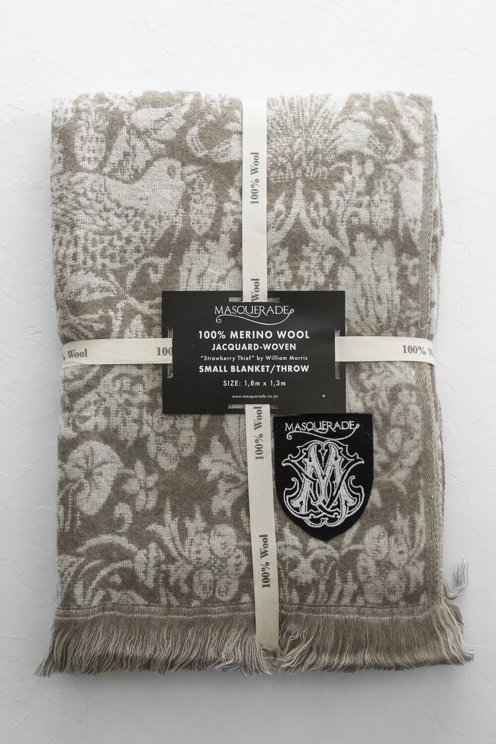 Beige jacquard-woven Merino wool blanket folded with fringe edge and Masquerade label ribbons on top.